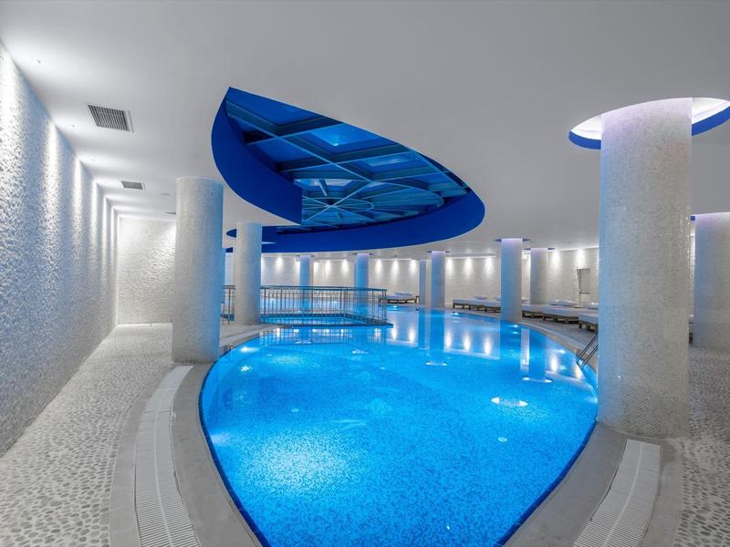 Innenraum mit beleuchtetem Schwimmbad, blauen Wasser, weißen Wänden und Säulen, modern gestaltet.