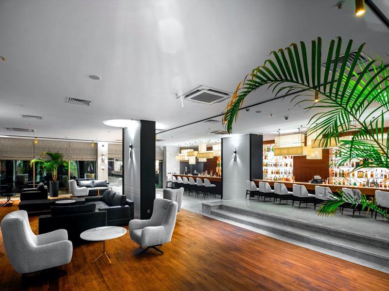 Moderner Hotel-Lobby mit Holzboden, grauen Sesseln, schwarzem Sofa und beleuchtetem Empfangstresen.