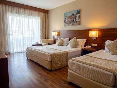 Helles Hotelzimmer mit Doppel- und Einzelbett, Holzboden und Balkon mit Vorhängen.