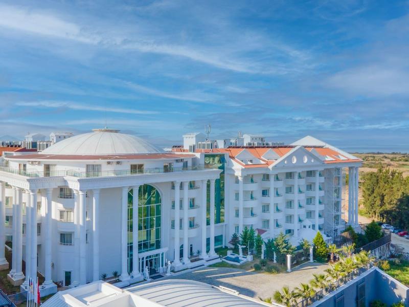 Grande hotel bianco con tetti rossi accanto a un'area boschiva sotto un cielo blu.