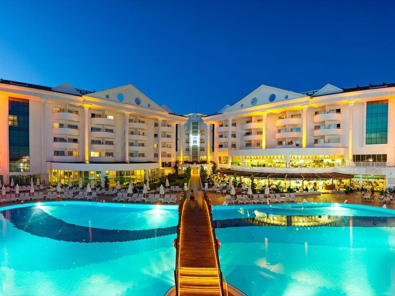 Grande hotel con piscine illuminate e passerella in legno al tramonto.