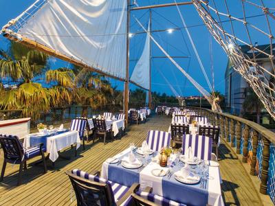 Terras van een restaurant op een zeilschip met gedekte tafels en uitzicht op palmbomen bij schemering.