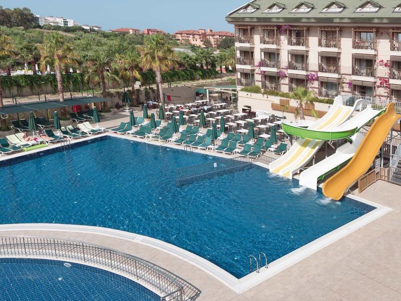 Duży basen hotelowy z zjeżdżalniami, leżakami i palmami obok wielopiętrowego budynku.