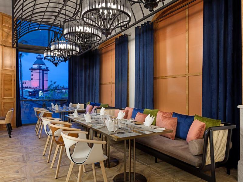 Elegancka jadalnia z dużymi oknami, kanapą z kolorowymi poduszkami i drewnianymi krzesłami.