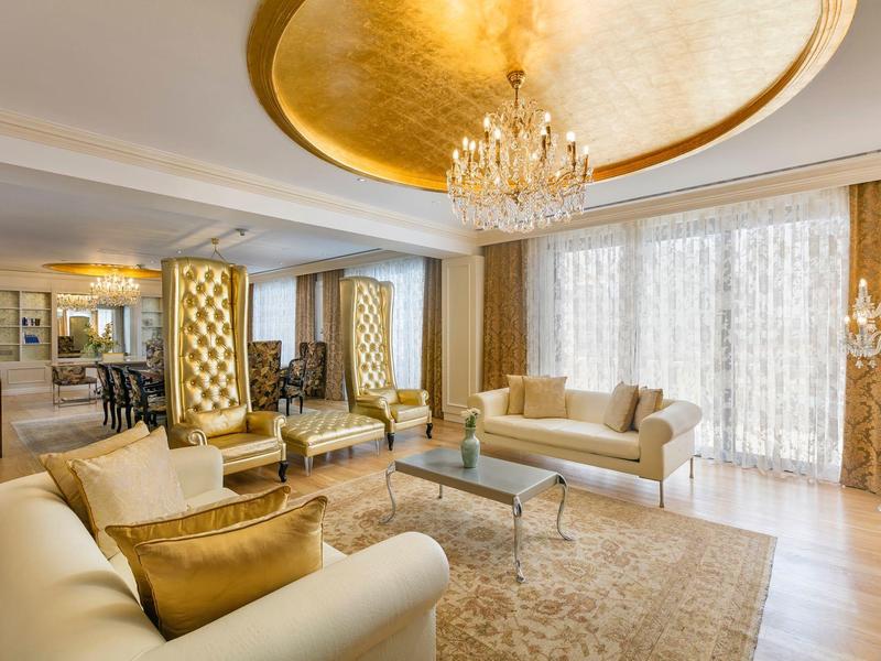 Elegancki salon hotelowy z beżową sofą, wysokim fotelem, złotym żyrandolem i dużymi oknami.