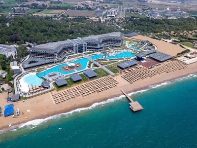 Großer Hotelkomplex mit Pools, Sandstrand und Steg am türkisblauen Meer, umgeben von grüner Landschaft.