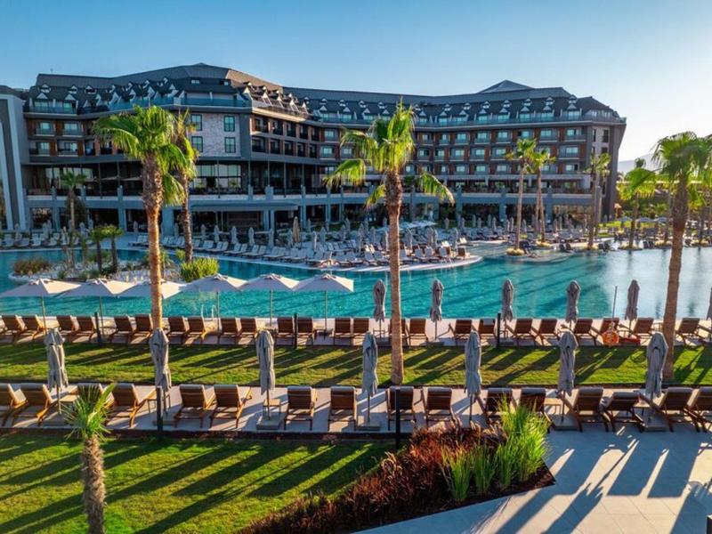 Duży hotel resort z basenem, palmami i leżakami o zachodzie słońca.