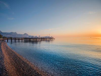 Peaceful beach with a wooden pier at sunrise over calm, clear water and pebble shore.