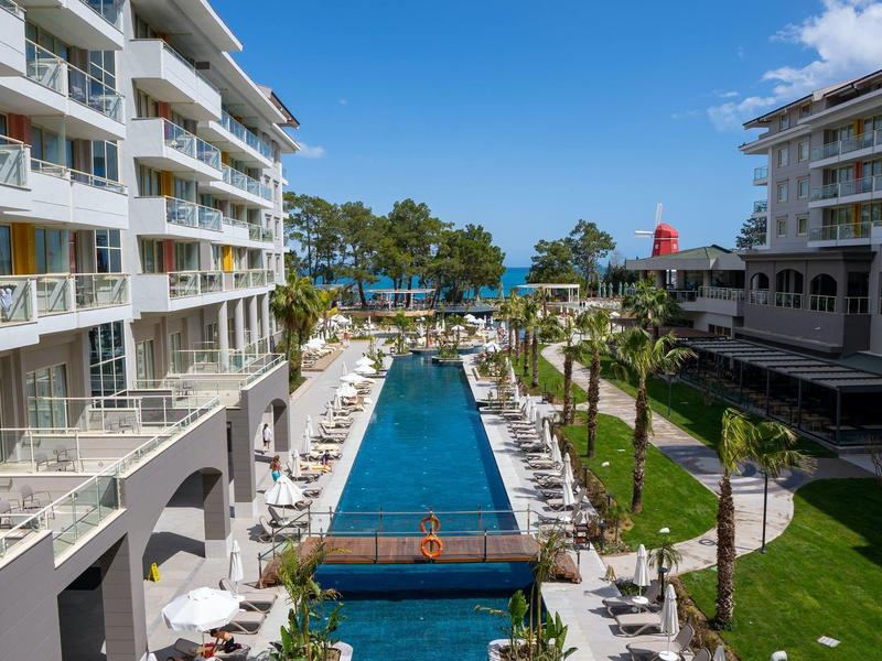 Outdoor pool with lounge chairs between two hotels, palm trees, and a sea view in the background.