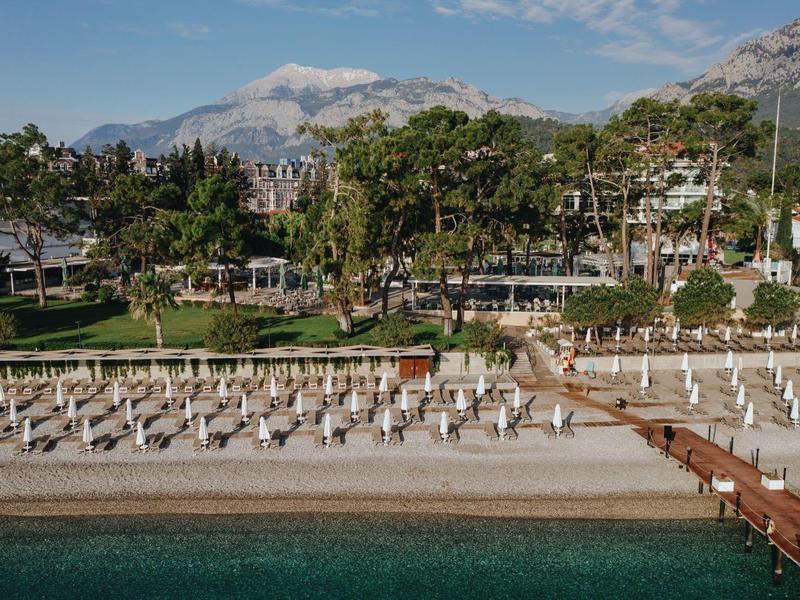Beachfront resort with rows of white lounge chairs, wooden pier, lush trees, and mountains in background.