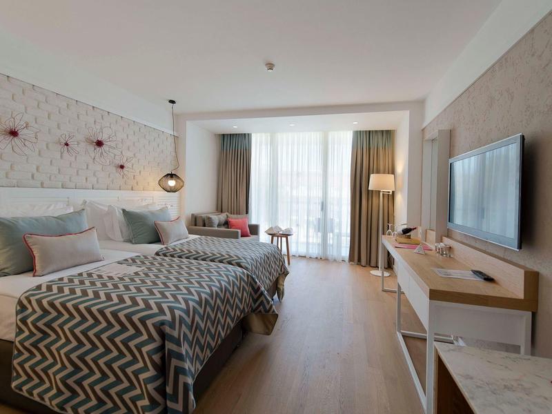 Bright hotel room with two beds, wooden floor, TV, and large window with curtains.