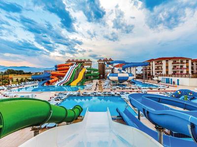 Velký vodní park s barevnými tobogány a bazénem před hotelovými budovami a horami.