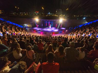 Grande pubblico assiste a uno spettacolo serale in un anfiteatro all'aperto illuminato.