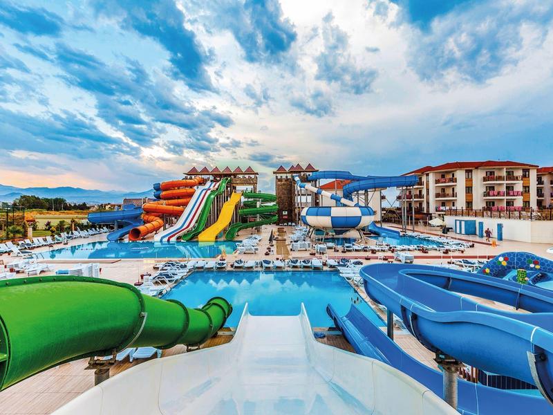 Velký vodní park s barevnými tobogány a bazénem před hotelovými budovami a horami.