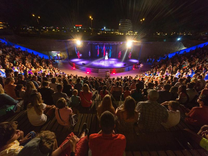 Plný amfiteátr během večerní akce s barevným osvětlením jeviště.