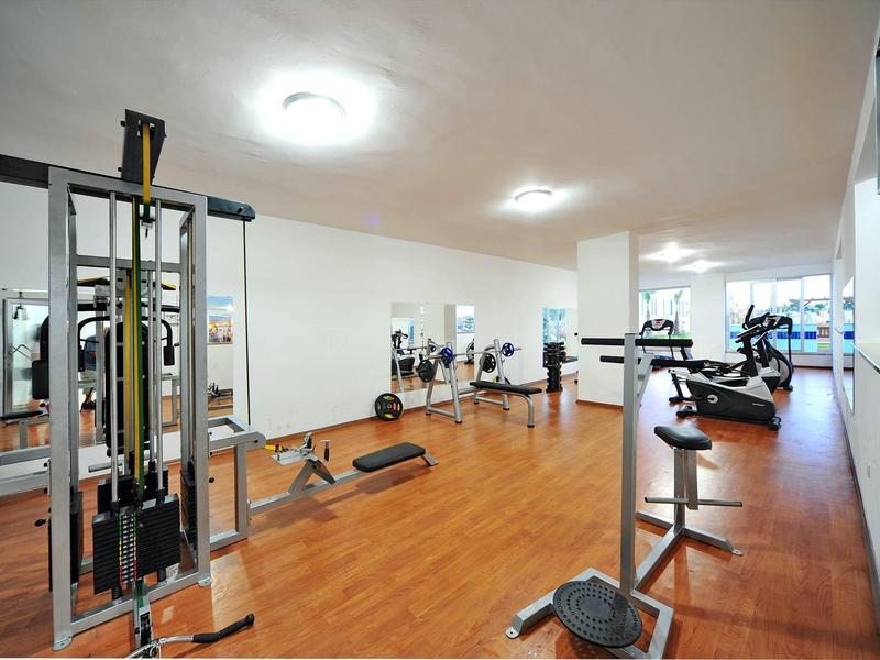 Moderní posilovna s činkami, cvičebními stroji a dřevěnou podlahou v světlé místnosti.