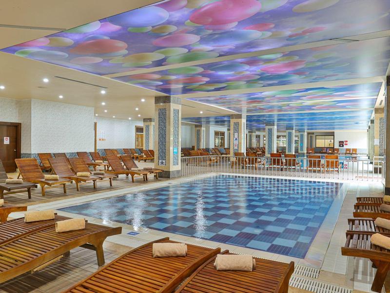 Área de piscina interior con tumbonas de madera y techo iluminado con colores en un hotel.