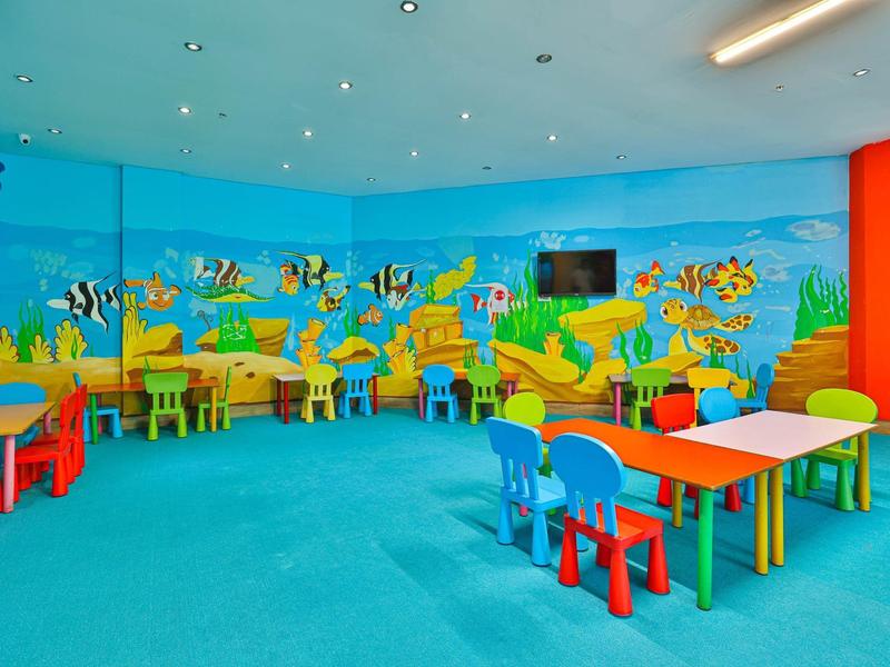 Sala de juegos colorida con mesas, sillas y pintura mural submarina en una zona de hotel.