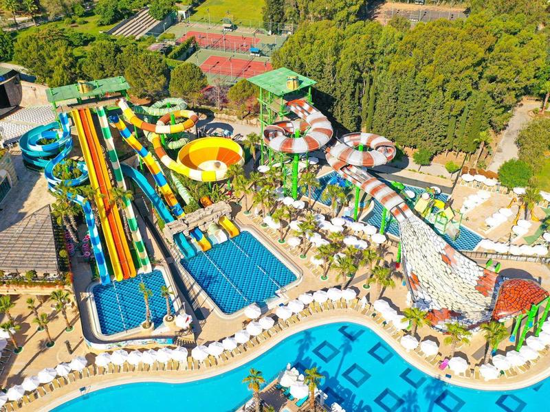 Parque acuático colorido con toboganes, piscinas y sombrillas rodeado de áreas verdes.