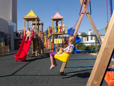 Kind schaukelt auf gelber Schaukel vor buntem Klettergerüst mit roten Rutschen auf Spielplatz.