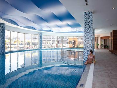 Hallenbad mit blauem Wellenmuster an der Decke, großen Fenstern und Frau am Beckenrand.