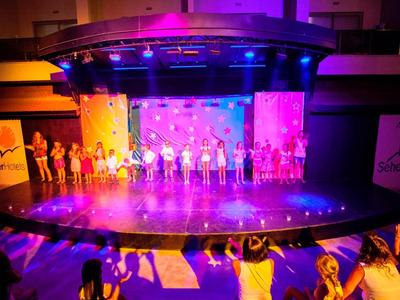 Bunt beleuchtete Bühne mit Menschen in Reihe, Publikum im Vordergrund, modernes Theaterambiente.