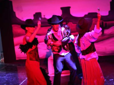 Drei Personen in Cowboy-Kostümen tanzen vor lila beleuchtetem Westernkulisse-Hintergrund.