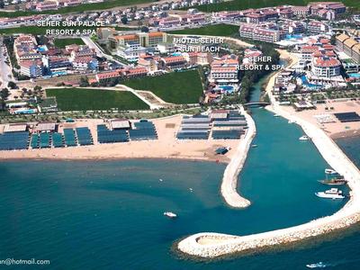 Luftaufnahme eines Strandes mit Sand, Pier, Booten und angrenzenden Häusern und Grünflächen.