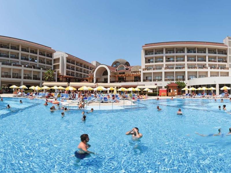 Large hotel pool with guests, surrounded by sun loungers and a modern multi-story hotel.