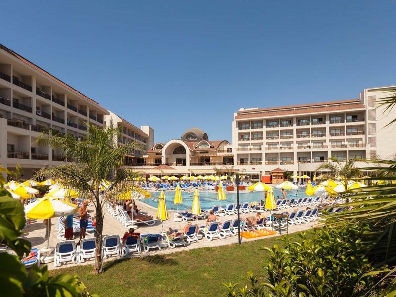 Large hotel with pool area, sun umbrellas, and lounge chairs on a sunny day.