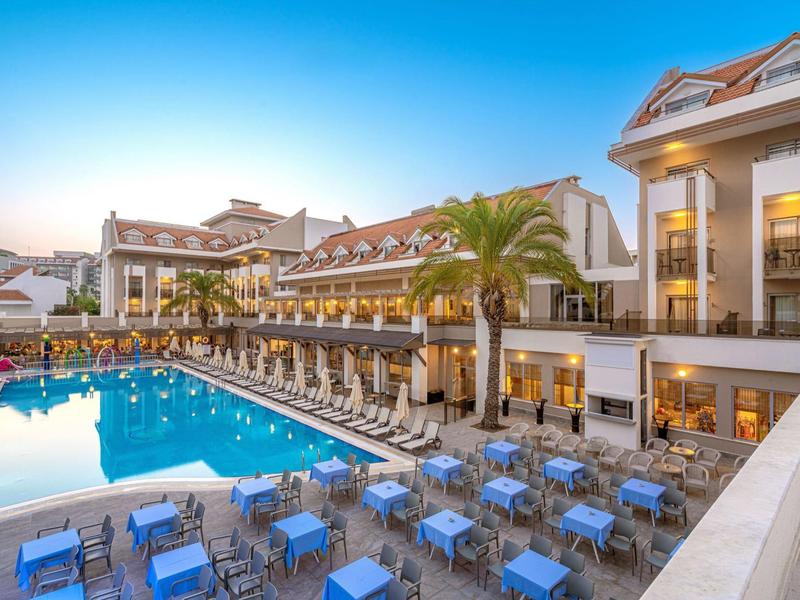 Hotel met groot buitenzwembad, veel tafels en parasols bij een heldere hemel.