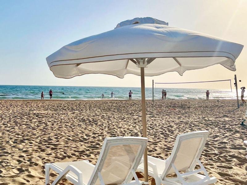 Twee witte ligstoelen onder een parasol op een zandstrand met de zee op de achtergrond.
