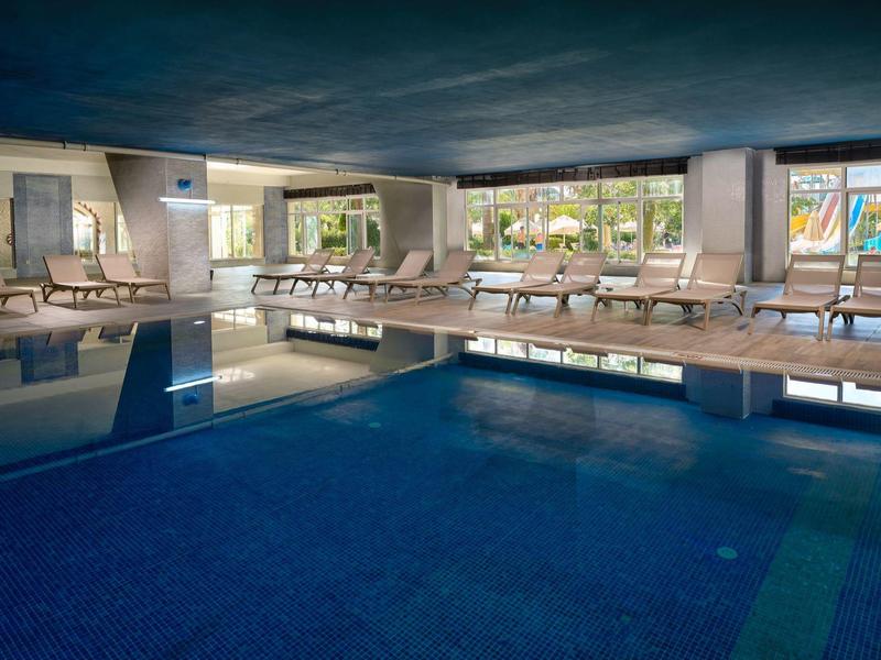 Espace piscine intérieur avec chaises longues et grandes fenêtres donnant sur l'extérieur dans un hôtel.