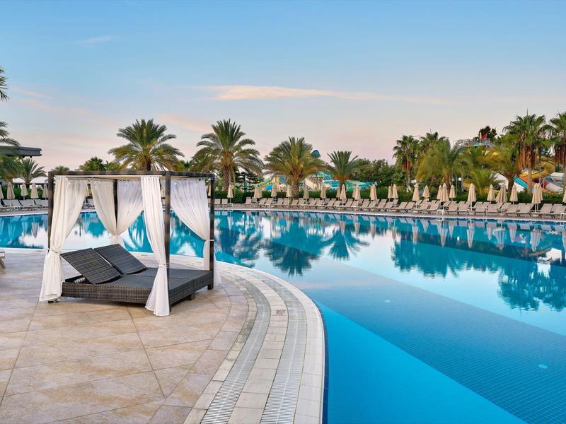 Grand piscine d'hôtel avec des palmiers et des chaises longues blanches à baldaquin au bord au coucher du soleil.