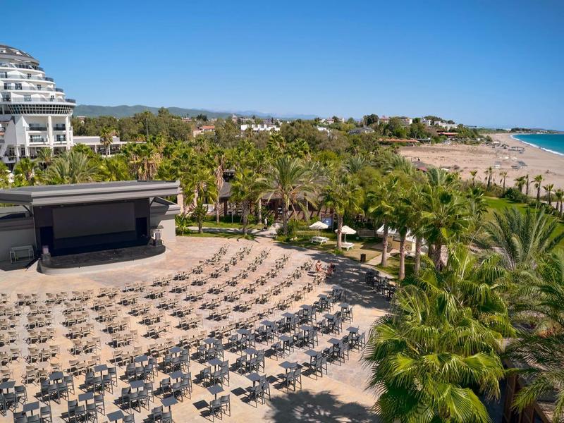 Complexe hôtelier avec scène, nombreuses chaises, palmiers et plage de sable au bord de la mer sous un ciel clair.