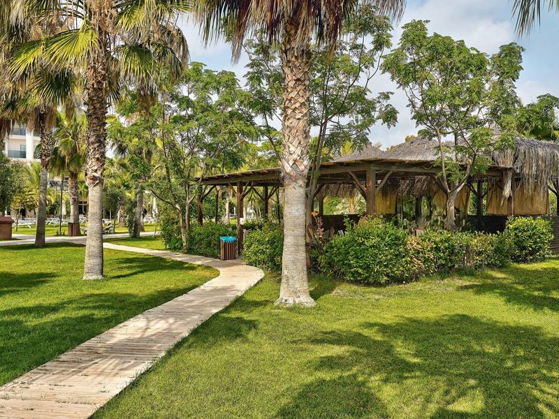 Complexe verdoyant avec palmiers, pelouse soignée et grand pavillon ouvert sous ciel bleu.