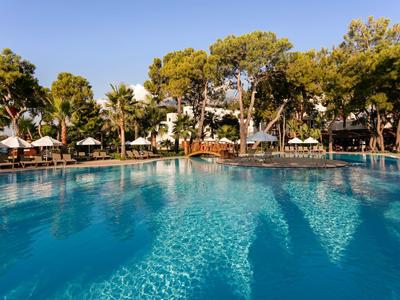 Großer blauer Pool mit Sonnenliegen, Sonnenschirmen und Bäumen im sonnigen Außenbereich.