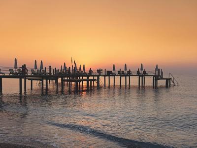 Langer Steg mit vielen geschlossenen Sonnenschirmen bei ruhigem Wasser im orangefarbenen Sonnenuntergang.