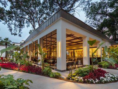Modernes Hotelrestaurant mit Glaswänden, umgeben von üppigen Gärten und Bäumen bei Dämmerung.