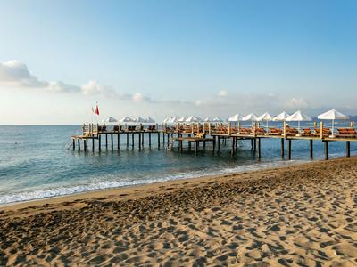 Ein langer Holzsteg mit Sonnenschirmen ragt ins ruhige Meer, mit sandigem Strand im Vordergrund.