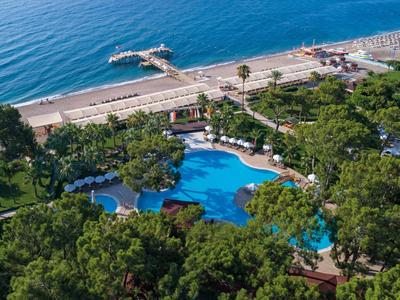 Großer blauer Pool umgeben von grünen Bäumen, nahe eines Sandstrands und blauen Meeres.