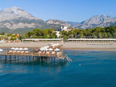 Klares blaues Meer mit Steg, Sandstrand, grünen Bäumen und hohen Bergen im Hintergrund.