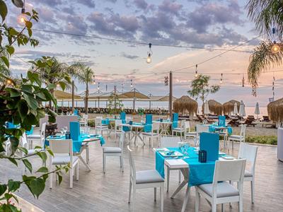 Strandrestaurant mit weißen Stühlen, blauen Tischdecken, Palmen und Wolkenhimmel bei Sonnenuntergang.