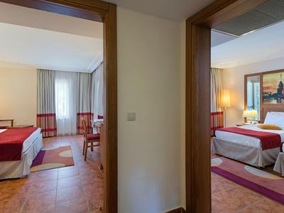 Innenansicht mit zwei angrenzenden Hotelzimmern, je mit Bett, Stuhl, Tisch und warmem Licht.