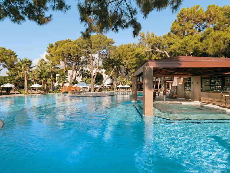 Großer blauer Pool mit Sonnenschirmen, Palmen und schattigem Pavillon im Hintergrund.