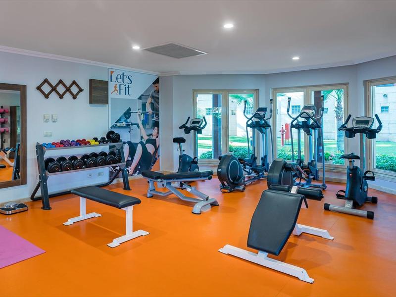 Moderner Fitnessraum mit orangem Boden, Geräten, Hanteln und großen Fenstern mit Blick ins Grüne.