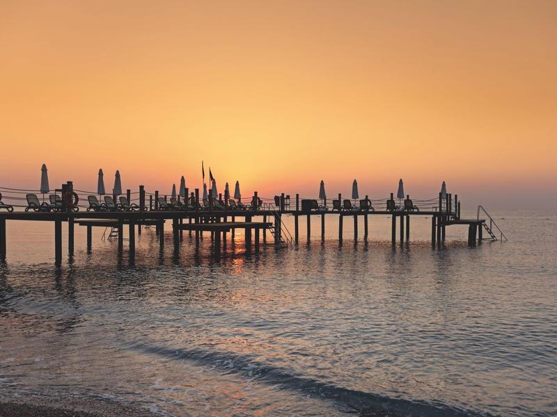 Langer Steg mit vielen geschlossenen Sonnenschirmen bei ruhigem Wasser im orangefarbenen Sonnenuntergang.