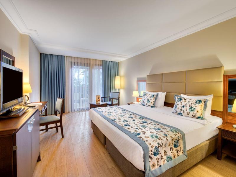 Hell eingerichtetes Hotelzimmer mit Doppelbett, TV, Schreibtisch und Balkon mit Vorhängen.