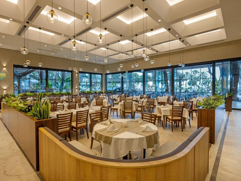 Helles Restaurant mit runden Tischen, weißen Tischdecken und großen Fenstern mit Blick auf die Natur.