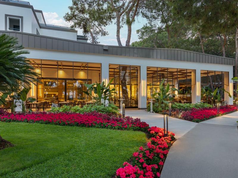 Hotelgebäude mit großen Fenstern, umgeben von grünen Bäumen und bunten Blumenbeeten.